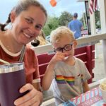 Photo of a woman (me) in an orange shirt, holding a purple travel mug with a straw sticking out of it, sitting on a red bench next to a little boy in a gray shirt (with a rainbow on it) teaching icecream.
