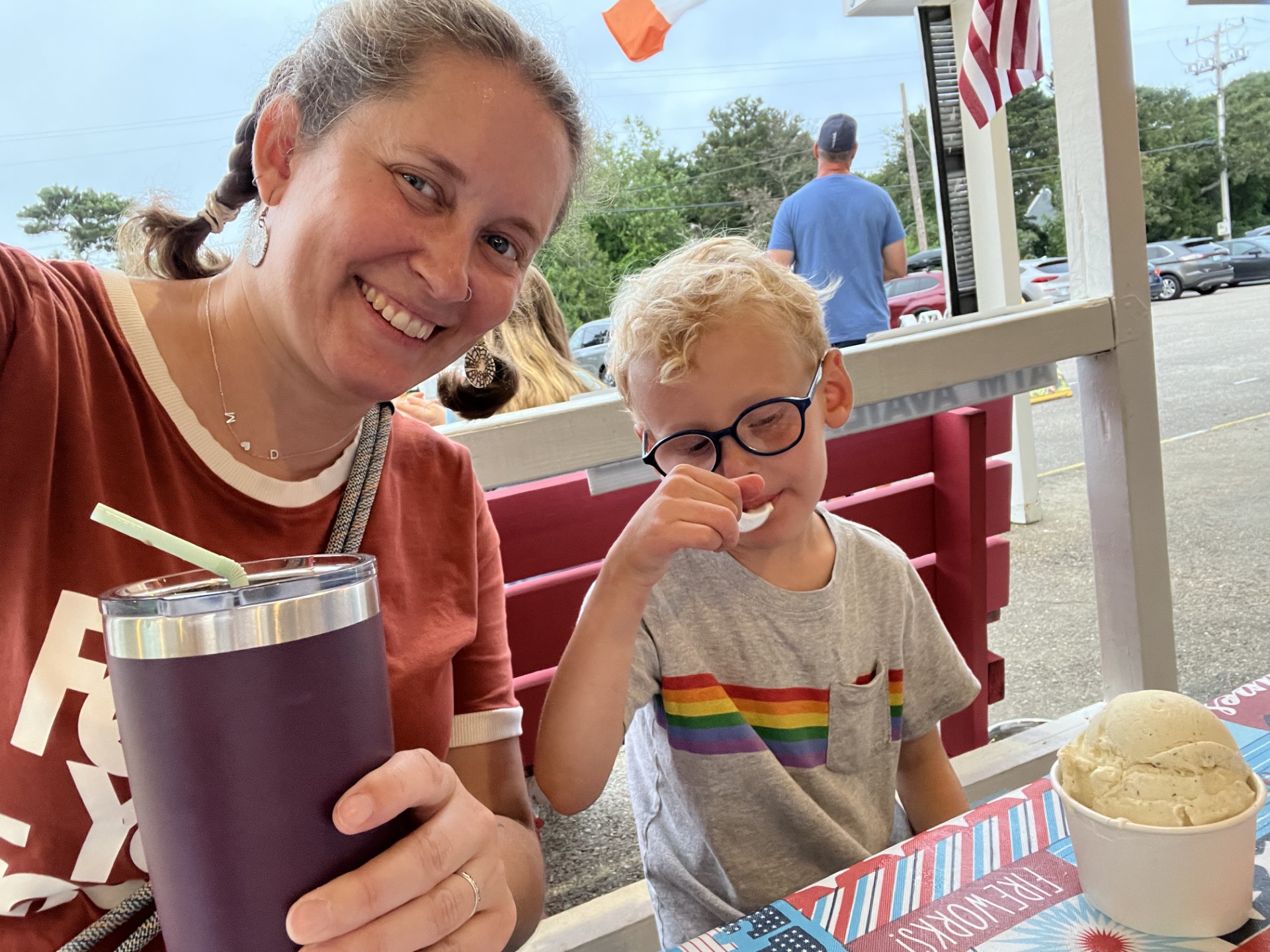 Photo of a woman (me) in an orange shirt, holding a purple travel mug with a straw sticking out of it, sitting on a red bench next to a little boy in a gray shirt (with a rainbow on it) and blue glasses eating ice cream.